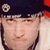 A pixelated close-up of a person wearing a dark cap with a circular emblem, light-colored shirt, and dark hair, with a red and white background.
