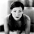 Pixelated grayscale image of a person with short dark hair wearing a dark sleeveless top, seated against a light background with a dark rectangular shape on the left.