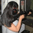 A person with long dark hair in a white sleeveless top holds a black rifle-like object, positioned against a blurred background with vertical elements.