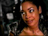 Pixelated close-up of a person with light brown skin tone, dark hair pulled back, and visible ear against dark background with indistinct shapes.