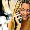 A woman with long blonde hair holds a dark green cell phone to her ear, smiling with closed eyes and visible teeth, wearing a light-colored sleeveless top and a beaded bracelet.