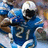 Pixelated image of a figure in blue sports uniform with number 2, white helmet, and brown gloves holding a football.