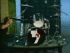 Animated sequence of a drummer playing drums with cymbals and drum kit, wearing black shirt, against green and blue background.