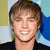 A person with light brown hair and blue eyes wears a black shirt against a background of yellow, white, and blue shapes.
