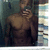 Pixelated static image of a shirtless figure with muscular build, hand near face, against dark and light background elements.
