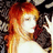 Pixelated image of a person with long red hair, closed eyes, and light skin wearing a white top against a dark background.