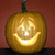 A pixelated pumpkin with a carved smiling face, glowing from within, set against a reddish-brown background.