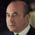 Pixelated close-up of a bald man in a dark suit and white shirt, facing left with a neutral expression against a dark background.