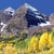 Pixelated landscape with rocky mountain peaks under blue sky, yellow and green foreground vegetation, and dark cave-like opening in rock face.