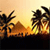 Pixelated sunset scene with palm trees silhouetted against warm orange and yellow sky, dark ground below.