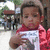 A pixelated image shows a child in red clothing holding a white sign with blue text, standing before an outdoor background with structures and greenery.