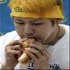 Static image of a person wearing a yellow cap and white shirt eating a sandwich with both hands, visible necklace, blue background.