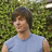 A pixelated image of a person with short brown hair wearing a light blue shirt, standing against a green leafy background.