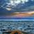 Pixelated landscape showing a body of water under a sky with clouds and a setting or rising sun, with a small landmass or structure in the foreground.