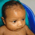 A baby with dark hair sits in a blue and teal tub, looking down with eyes partially closed.