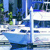 Pixelated static image of a white boat with blue hull docked at a marina, surrounded by water and greenery under a blue sky.