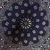 A static digital icon featuring a symmetrical floral or mandala design with dark blue petals and light gray circular accents on a textured gray background.