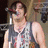 A person in a sleeveless patterned vest stands before a microphone, with a blurred background showing stage equipment.