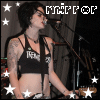 A person with tattoos and short dark hair sings into a microphone, wearing a black crop top with white text, surrounded by white stars on a dark background.