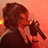 Pixelated image of a person with dark hair holding a microphone against an orange background, appearing to sing or speak.
