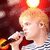 A person with light brown hair holds a microphone, wearing a striped shirt against a red and yellow background with a black text banner.