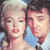 Pixelated image of two people in close proximity, one with light-colored hair and the other with dark hair, both facing forward against a blue and white background.