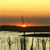 Sunset over water with a dark silhouette of a structure on the horizon, transitioning from yellow to orange to brown sky and reflecting in the water below.