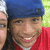 Two people in close-up; one wears a blue cap and red shirt, the other has long dark hair. Background is greenish-yellow.