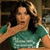 A pixelated image of a person with dark hair wearing a teal top, holding an object near their face against a blurred background.