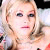 Pixelated close-up of a person with light-colored hair, fair skin, and dark eyes, wearing a light-colored top with visible shoulder straps.