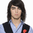 Pixelated portrait of a person with long dark hair wearing light blue shirt and black vest, red circular symbol on left chest.