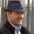Pixelated image of a person wearing a dark fedora, dark jacket, and light blue striped shirt, standing against vertical metal bars.