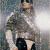 Pixelated static image of a person in a light-colored jacket and dark pants standing against a blurred background with muted tones.