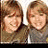Two pairs of people with brown hair pose back-to-back, one pair in gray and dark clothing against white background, the other in light and dark clothing against pink background.