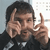 A person in a dark jacket and checkered shirt makes a two-finger gesture with both hands, first with the left hand raised higher than the right, then with both hands at equal height.
