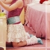 A person in a maroon sleeveless top and white polka-dot skirt kneels on a pink floor beside a light wooden table with a green bowl.