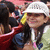 A person wearing a white cap with text, a light green jacket, and a pink lanyard, smiling in a crowd with red and blue clothing visible.