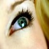 Close-up of a human eye with green iris, black eyelashes, and visible redness near the inner corner, set against light skin and a blurred background.