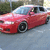 Pixelated image of a red sedan with light blue roof parked on gray pavement near green foliage.