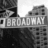 Black-and-white cityscape with 'BROADWAY' street sign and yellow pixelated text on black background reading 'One day you'll see my name up there in lights'.