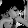 Black-and-white photo of a person with short dark hair resting their chin on a hand, wearing a sleeveless top with a visible tattoo on the shoulder.
