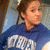 Pixelated image of a person with brown hair tied back, wearing a blue shirt with white text on it, against a blurred background.