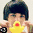 A person with dark hair holds a yellow rubber duck in front of their face, then covers one eye with a white mask, and finally holds a light blue heart-shaped object.