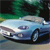 Pixelated image of a light blue sedan with dark blue accents on a road, green foliage in the background, and a blurred sky.