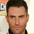 A static image of a person with short dark hair and light skin, wearing a black shirt against an orange background.