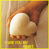 A hand holds a white heart-shaped object against a wooden background with yellow text reading "I GIVE YOU MY HEART." and "PLEASE DON'T BREAK IT.".