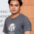 A person wearing a dark gray t-shirt with white stylized text on the chest, standing against an orange and light gray background.