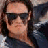 Pixelated close-up of a person with dark hair wearing dark sunglasses and a dark jacket against a blurred background.