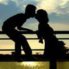 Silhouette of two figures leaning toward each other in a kiss, positioned on a railing with a sunset sky and water reflection below.