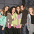 Six people stand closely together in a group pose, wearing varied casual clothing against a dark background.